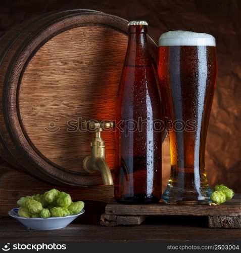 Bottled and unbottled beer glass with vintage old barrel and fresh hops for brewing still-life
