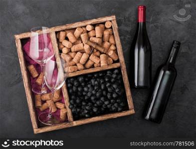 Bottles of red wine on wood with empty glasses and dark grapes with corks and corkscrew inside vintage wooden box on dark wooden background with red cloth.
