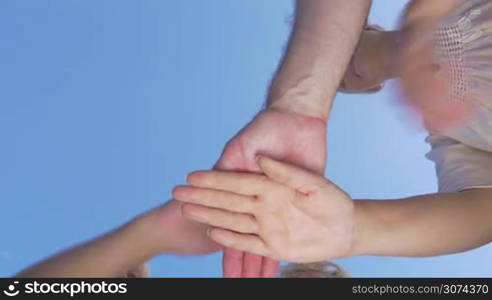 Bottom shot of young and senior people putting hands together and raising them on blue sky background. Motivation and teamwork