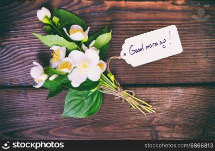 bouquet of blossoming jasmine with white flowers and a paper tag with the inscription good morning on a brown dock background