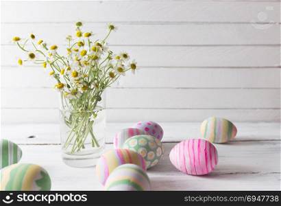 Bouquet of chamomiles in a glass vase and easter eggs in pastel color on white vintage planks