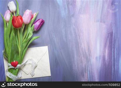 Bouquet of colorful tulips tied with a lace ribbon and a wooden clip with a red heart, and a closed envelope, on a purple wooden background.