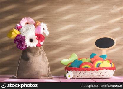 Bouquet of diverse flowers in a jute sack, a basket full of colorful and multi-shaped cookies, with a blank banner, on a pink table, on a sunny day.