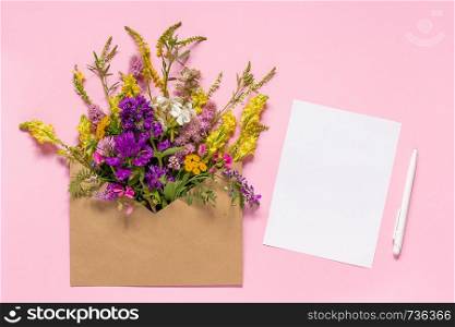 Bouquet of field multicolored flowers in craft envelope and white empty paper card for text with pen on pink background. Greeting card Flat Lay Mock-up Concept Women's day or Mothers Day.. Field flowers in craft envelope and white empty paper card