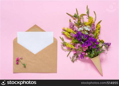 Bouquet of field multicolored flowers in waffle cone and craft envelope with white empty paper card for text on pink background. Greeting card Flat Lay Mock up Concept Women's day or Mothers Day.. Bouquet flowers in waffle cone and craft envelope with paper card