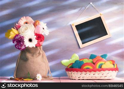 Bouquet of flowers in a jute sack, a basket full with cheerful multi-shape cookies and a blank blackboard on a purple wall, with light leaks.