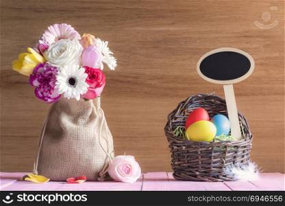Bouquet of flowers in a jute sack and a wicker basket with painted eggs and a blank wooden banner, on a pink table, with a wooden wall.