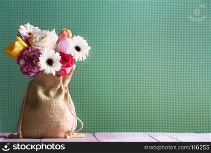Bouquet of multicolored gerbera, tulips, and roses in a cute hessian sack, on a pink wooden table, and a green fabric background, in natural light.