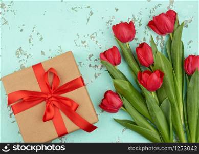 bouquet of red blooming tulips with green leaves, wrapped gift in brown craft paper and tied with a silk red ribbon on a green background, top view. Festive backdrop for birthday, Valentine&rsquo;s day
