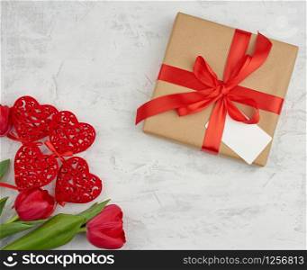 bouquet of red blooming tulips with green leaves, wrapped gift in brown craft paper on a white background. Festive backdrop for birthday, Valentine&rsquo;s day