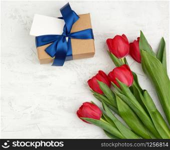 bouquet of red blooming tulips with green leaves, wrapped gift in brown craft paper on a white background, copy space. Festive backdrop for birthday, Valentine&rsquo;s day