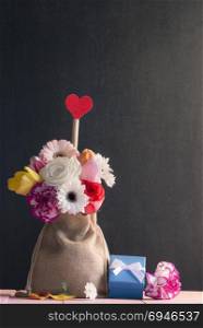 Bouquet of roses, gerbera, tulips and a red heart, in a jute bag, surrounded flowers and a cute blue gift box, with a black vintage background.