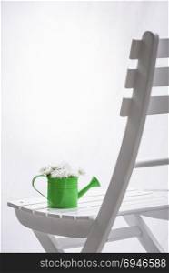 Bouquet of white delicate daisies in a green watering can on a white wooden chair, in front of a window with white curtains.