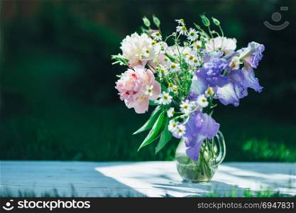 Bouquet of white peonies, chamomiles and iris flowers in glass vase. Summer background. Tinted photo. Bouquet of white peonies, chamomiles and iris flowers in glass vase. Summer background. Tinted photo.