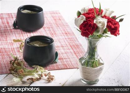 Bouquet of white roses and red carnations in a cute vase and two black cups of tea with dried linden flowers, on a white table, in sunlight.