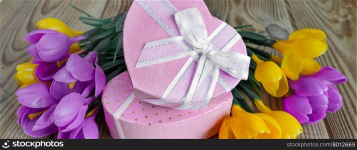 Bouquet of yellow and purple crocuses and two pink gift boxes in the shape of a heart, tied with white ribbons, on an unpainted wooden surface