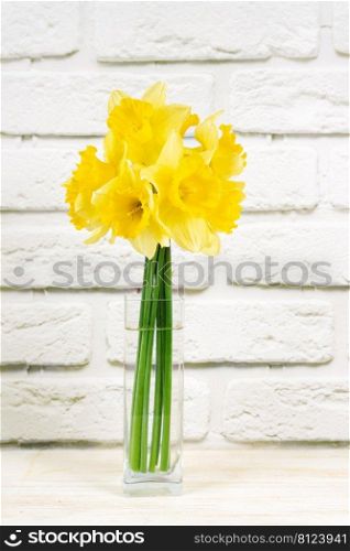 Bouquet of yellow daffodils, narcissus in transparent vase on white brick background with copy space. Mockup, template for holiday, birthday, mother’s day on yellow background with copy space for text. Bouquet of yellow daffodils, narcissus in vase on white brick background with copy space