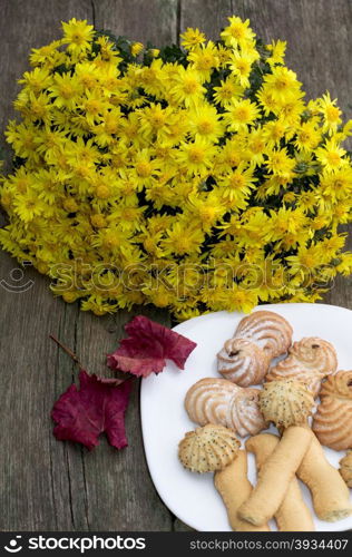 bouquet of yellow flowers, red leaf and plate with cookies, on a wooden table, fall a still life, a subject of sweet and flowers