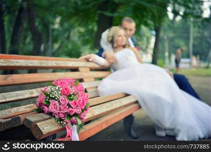 bouquet on wooden bench with bride and groom in the background, focus on the flowers. flowers on the background of the newlyweds. bouquet on wooden bench with bride and groom in the background, focus on the flowers