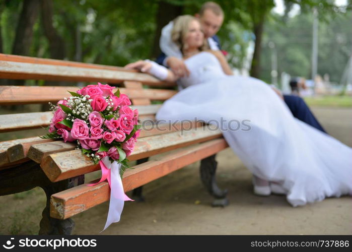 bouquet on wooden bench with bride and groom in the background, focus on the flowers. flowers on the background of the newlyweds. bouquet on wooden bench with bride and groom in the background, focus on the flowers