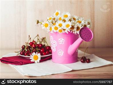 Bouquets of daisies inside rustic watering pot, on a white napkin and wooden background. Alongside is a plate full of cherries