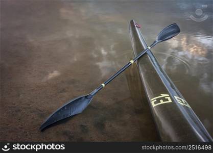 bow of narrow racing kayak with a wing paddle on a shore of calm lake at sunset, thirteen - temporary race number placed on deck by myself