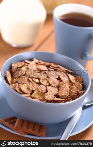 Bowl of chocolate corn flakes cereal with spoon and chocolate bars, a cup of tea and a jug of milk in the back (Selective Focus, Focus in the middle of the cereal). Chocolate Corn Flakes
