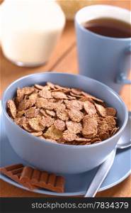 Bowl of chocolate corn flakes cereal with spoon and chocolate bars, a cup of tea and a jug of milk in the back (Selective Focus, Focus in the middle of the cereal). Chocolate Corn Flakes
