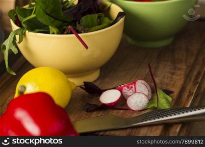 Bowl of fresh herbs with rocket and baby spinach and assorted salad ingredients including a lemon, red bell pepper and radish on a kitchen counter waiting to be prepared for dinner