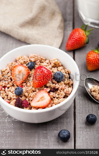 Bowl of healthy cereal granola with strawberries and blueberries and glass of milk on wooden board