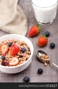 Bowl of healthy cereal granola with strawberries and blueberries and glass of milk on wooden board