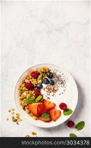 Bowl of homemade granola with yogurt and fresh berries on white marble background. Top view, flat lay. Healthy breakfast concept. Space for text. Bowl of homemade granola with yogurt and fresh berries. Bowl of homemade granola with yogurt and fresh berries