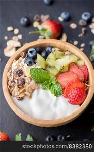 bowl of oat granola with yogurt, fresh blueberries, strawberries, kiwi mint and nuts board for healthy breakfast,  Healthy breakfast menu concept. On the black rock