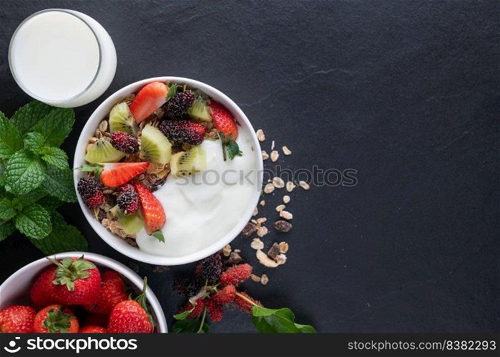 bowl of oat granola with yogurt, fresh mulberry, strawberries, kiwi mint and nuts on the black rock board for healthy breakfast, top view, copy space, flat lay. Healthy breakfast menu concept.
