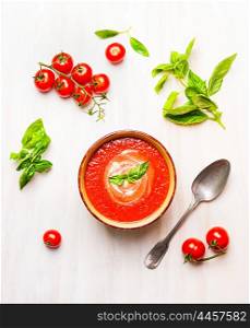 Bowl of tomato soup or gazpacho with spoon and basil on white wooden background, top view