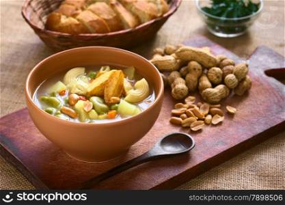 Bowl of traditional Bolivian Sopa de Mani (peanut soup) made of meat, pasta, vegetables (pea, carrot, potato, broad bean, pepper, corn) and ground peanut, photographed on wooden board with natural light (Selective Focus, Focus in the middle of the soup)