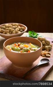Bowl of traditional Bolivian Sopa de Mani (peanut soup) made of meat, pasta, vegetables (pea, carrot, potato, broad bean, pepper, corn) and ground peanut, photographed on wooden board with natural light (Selective Focus, Focus one third into the soup)