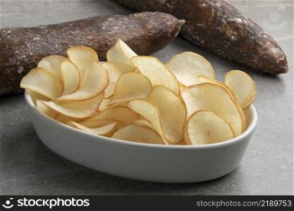 Bowl with deep fried cassava chips close up and waxed cassave tuber in the background