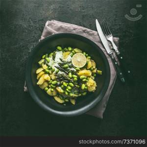 Bowl with potatoes salad with roasted green asparagus, edamame soybeans, lime and green peas on dark rustic kitchen table background, top view. Copy space. Healthy vegetarian food .