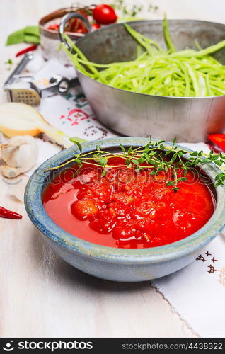 Bowl with tomatoes sauce on kitchen table with cooking pot and green vegetables, close up