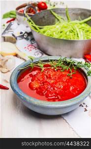 Bowl with tomatoes sauce on kitchen table with cooking pot and green vegetables, close up
