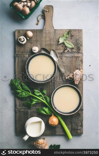 Bowls of homemade mushroom soup and ingredients on grey concrete background, top view