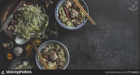 Bowls with Asian soup with cabbage, mushrooms, shiitake, garlic, chili and dumpling in traditional crockery with chopsticks on dark concrete table. Food banner with healthy ramen noodles. Top view