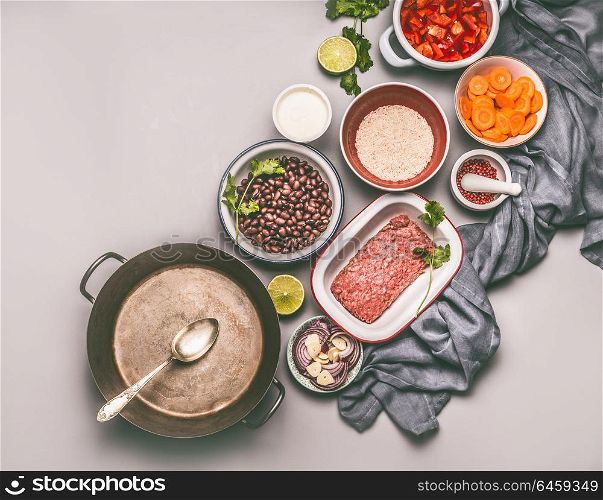 Bowls with ingredients for balanced one pan meal with beans, minced meat, rice and various cut vegetables and cooking pot with spoon on gray background, top view