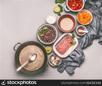 Bowls with ingredients for balanced one pan meal with beans, minced meat, rice and various cut vegetables and cooking pot with spoon on gray background, top view