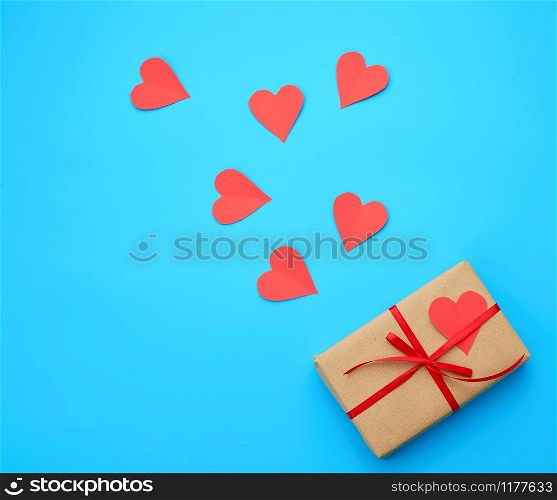 box is wrapped in brown paper and tied with a red thin silk ribbon on a blue background, red cut out paper hearts, copy space, holiday is Valentine&rsquo;s Day
