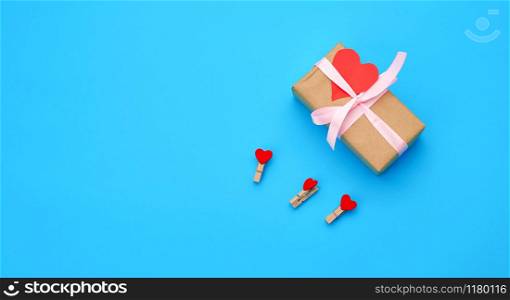 box is wrapped in brown paper and tied with a red thin silk ribbon on a blue background, red cut out paper hearts, copy space, holiday is Valentine&rsquo;s Day