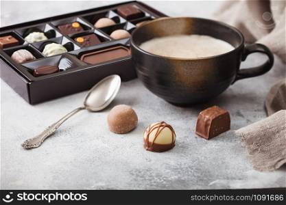 Box of Luxury Chocolate candies selection with cup of cappuccino coffee and silver spoon on light background.