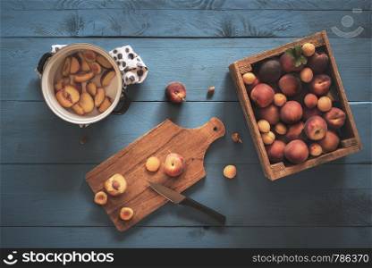 Box with fresh peaches and apricots, slicing fruits on cutting board and pot with fruit pieces for making jam. Above view with home making jam.