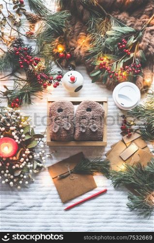 Box with funny handmade teddy-bear socks on white blanket with winter decorations, burning candles, pine branches, fairy lights and kraft paper envelope. Christmas gifts wrapping preparation.